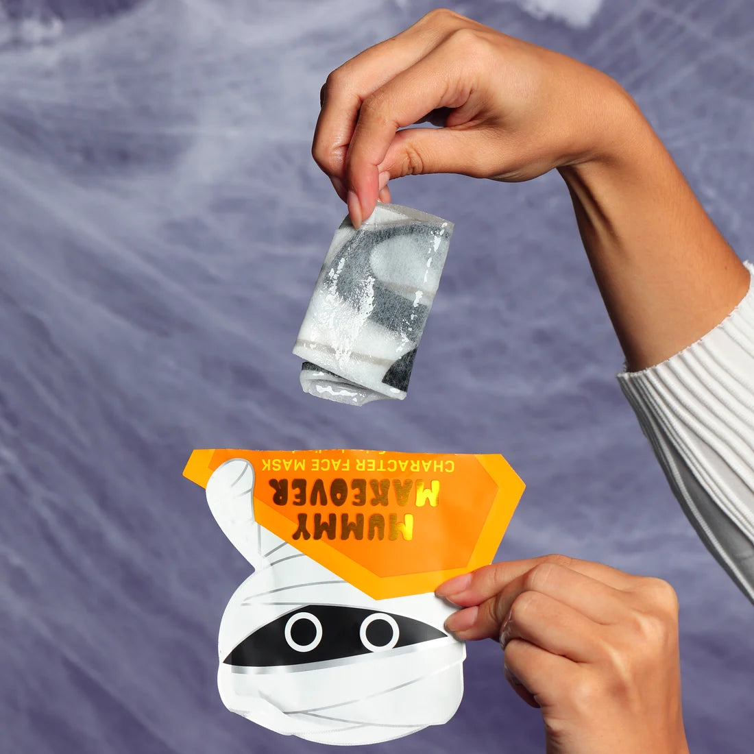 Mummy character facial mask sachet with illustrated face, held by hands